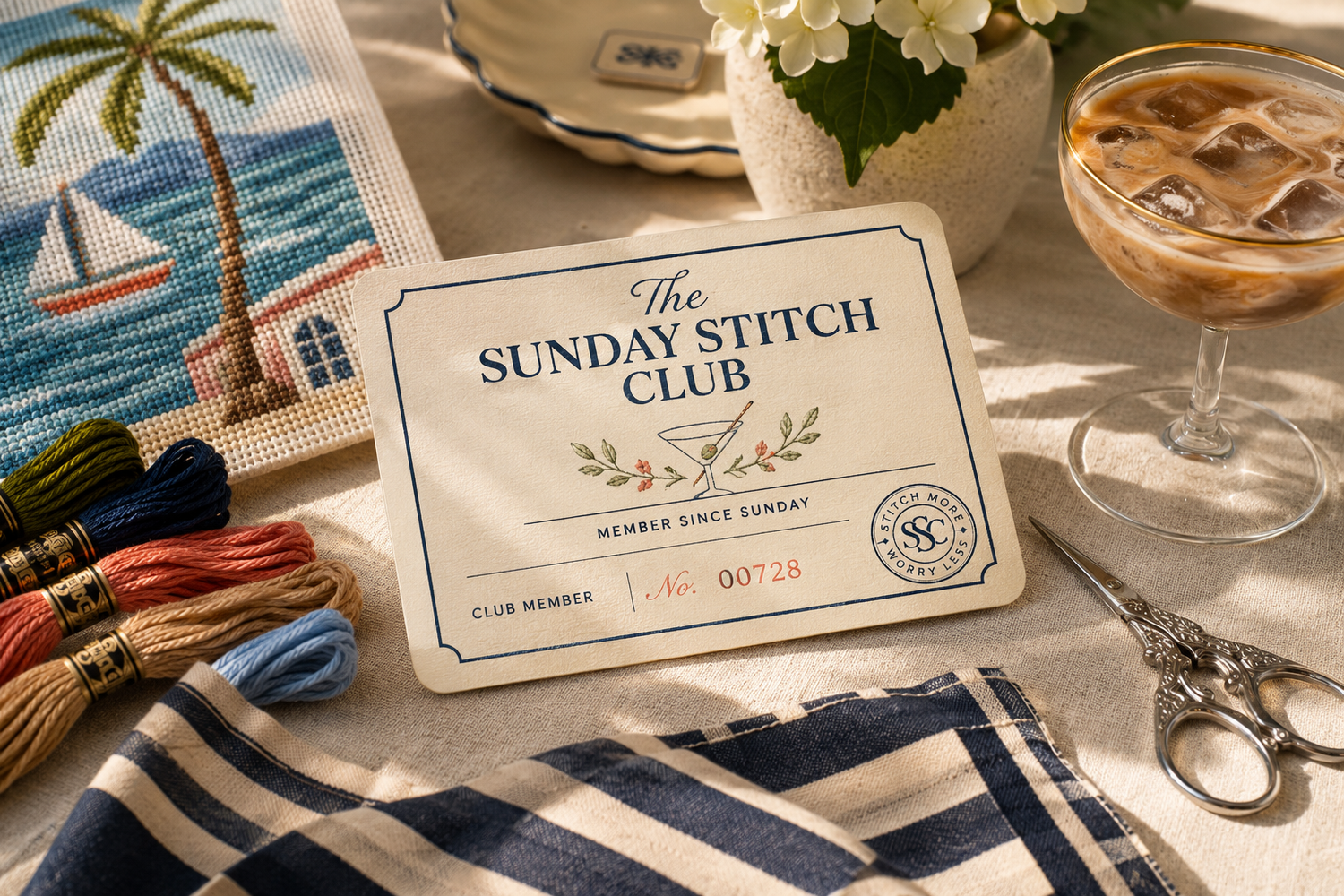 The Sunday Stitch Club card on a table with needlepoint materials and a drink.