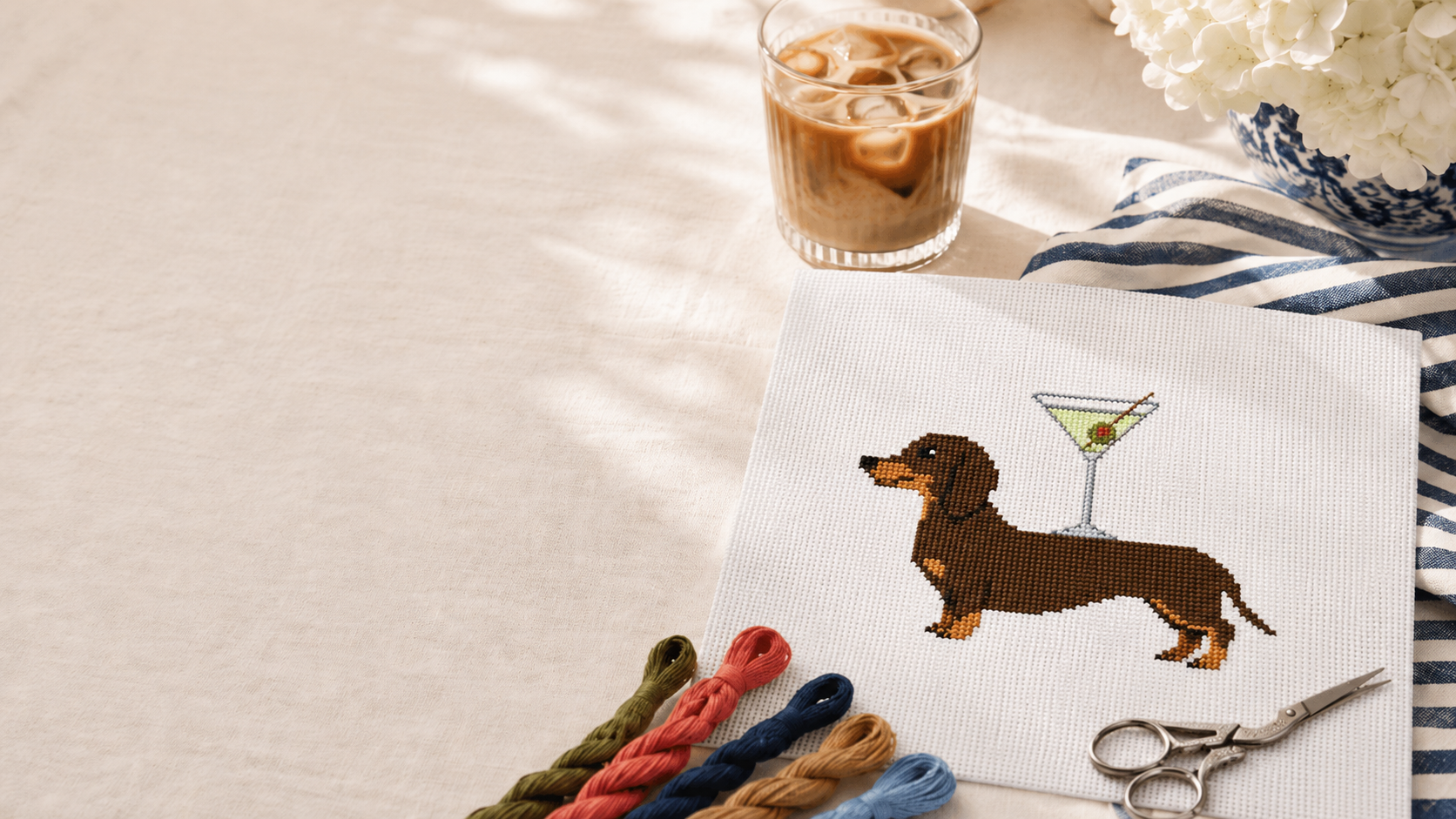 Cross-stitched dachshund with a martini glass on fabric with yarn and scissors.